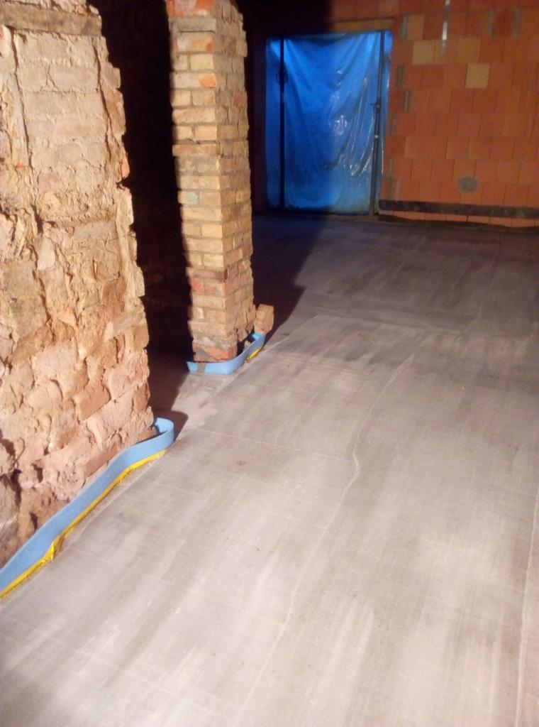 Świeżo wylany betonowy fundament wewnątrz budynku, zabezpieczony niebiesko-żółtą taśmą przy ceglanych filarach i ścianach z pustaków, z zakrytym folią wejściem w tle.