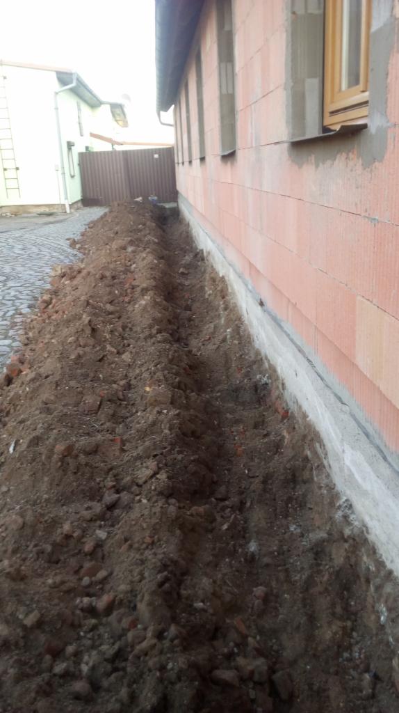 Wykopany wąski rów wzdłuż ściany budynku z czerwonej cegły, widoczny fragment fundamentu, na górze kostka brukowa i ogrodzenie.