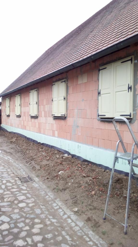 Budynek z czerwonej cegły z oknami z okiennicami, izolowany fundament, widoczna drabina i brukowa droga.