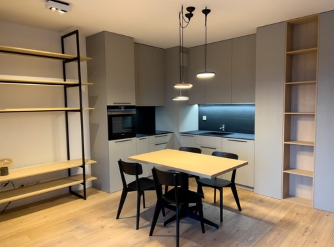 Nowoczesna, minimalistyczna kuchnia z szarymi szafkami, drewnianym blatem stołu i czarnymi krzesłami, oświetlona trzema wiszącymi lampami. Po lewej stronie regał z drewnianymi półkami i czarną ramą...