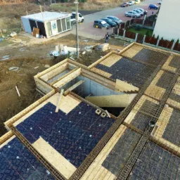 Widok z góry na budowę fundamentów domu z widocznym zbrojeniem, szalunkiem i częściowo ułożonymi płytami, z żółtą koparką w tle i budynkiem biurowym w oddali.