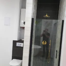 Nowoczesna łazienka z kabiną prysznicową typu walk-in, złotą armaturą, wiszącą toaletą i wzorzystą podłogą, sfotografowana z widocznym odbiciem fotografa w szklanej ściance.