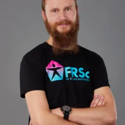 Portret brodatego mężczyzny z włosami związanymi w kitkę, ubranego w czarną koszulkę z kolorowym logo, stojącego na szarym tle z założonymi rękami.