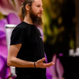 Mężczyzna z brodą w czarnej koszulce gestykuluje dłonią, w tle rozmyte kwiaty i elementy wyposażenia gabinetu.