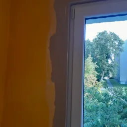 Białe okno z widokiem na zieleń, ściana w kolorze pomarańczowym w trakcie malowania, widoczne ślady szpachli przy ościeżnicy.