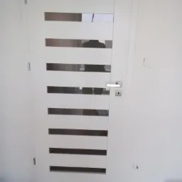 Białe drzwi wewnętrzne z poziomymi przeszkleniami, widoczna klamka i zamek, ściana z widocznymi śladami po instalacji.