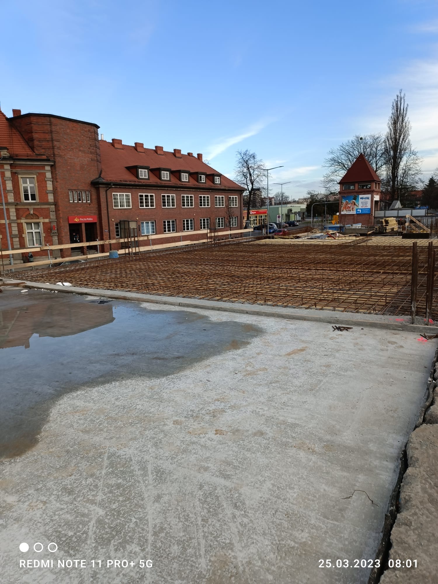Przygotowanie zbrojenia na budowie w Zabrzu. Widoczna siatka z prętów stalowych na betonowej płycie, w tle budynek Poczty Polskiej i wieża ciśnień.