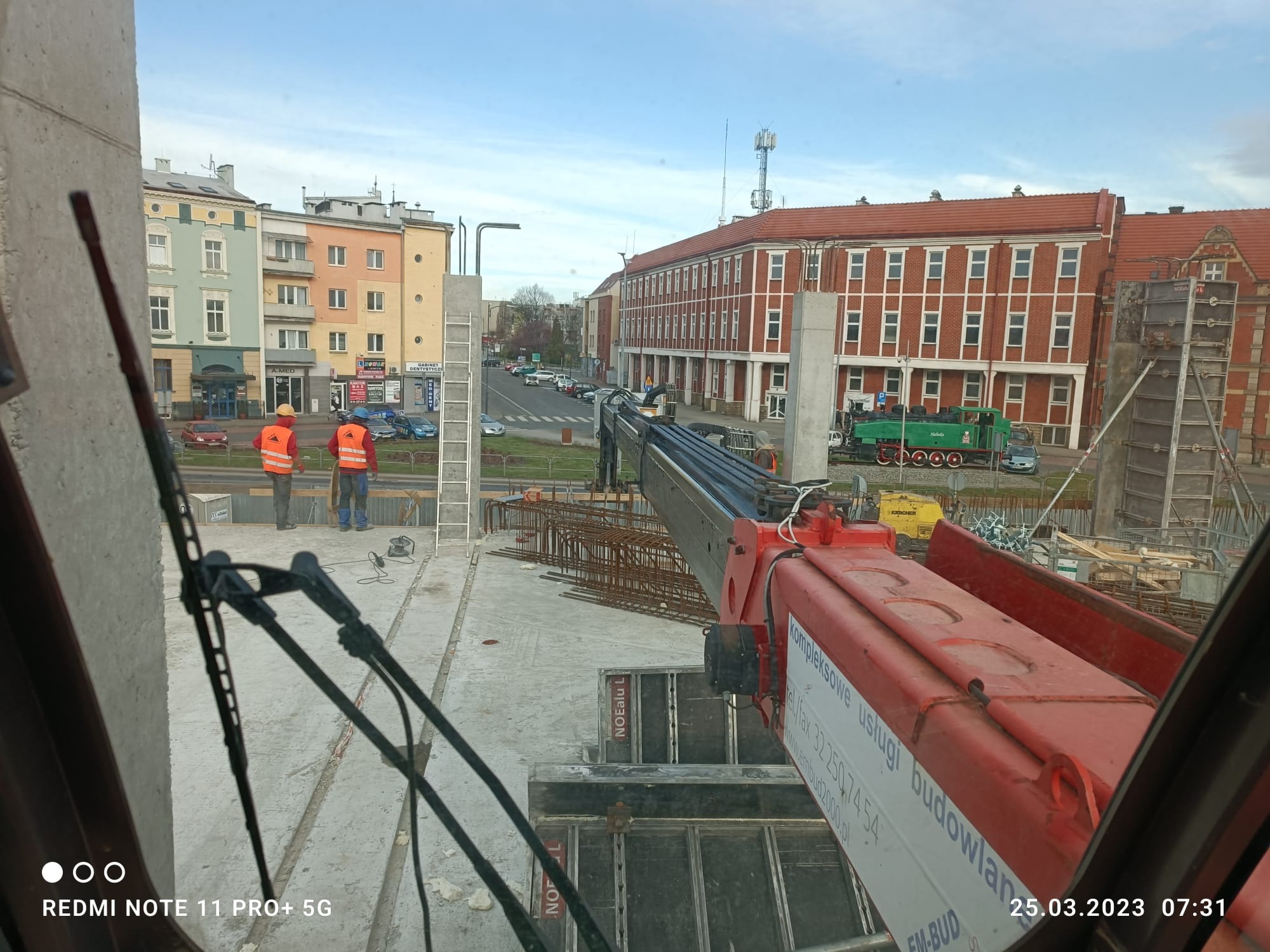 Budowa Dworca PKP w Raciborzu w trakcie robót żelbetowych: widok z kabiny dźwigu na pracowników w pomarańczowych kamizelkach, zbrojenie, szalunki NOEalu L i fragmenty dźwigu z napisem 'kompleksowe...