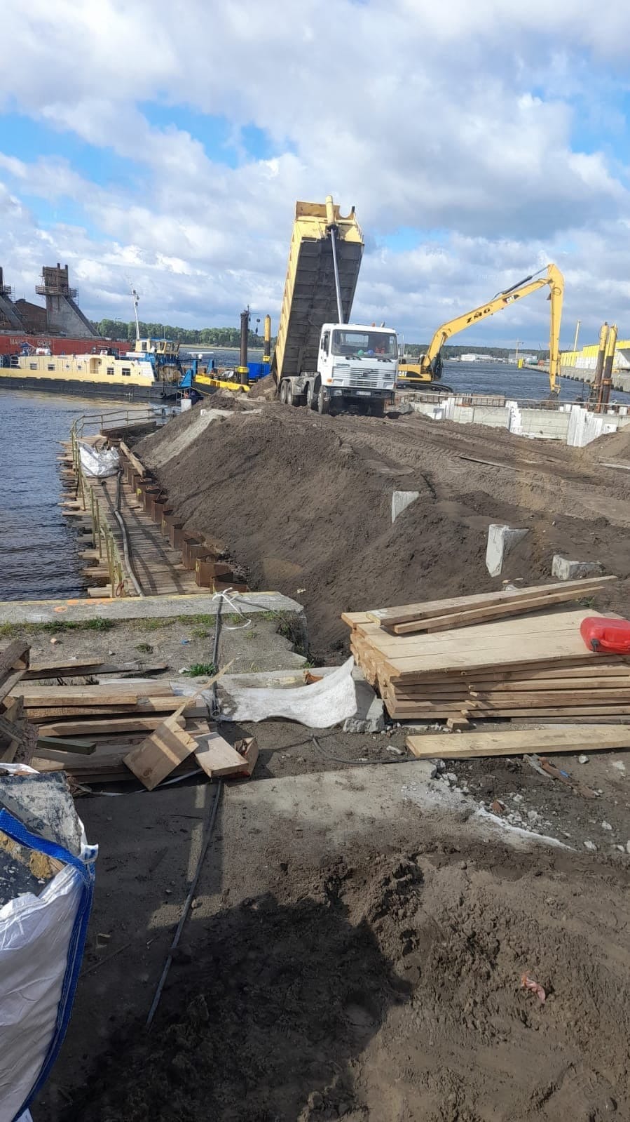 Budowa nabrzeża: wywrotka wysypuje ziemię na przygotowany teren. W tle widoczny dźwig i elementy konstrukcyjne nabrzeża.