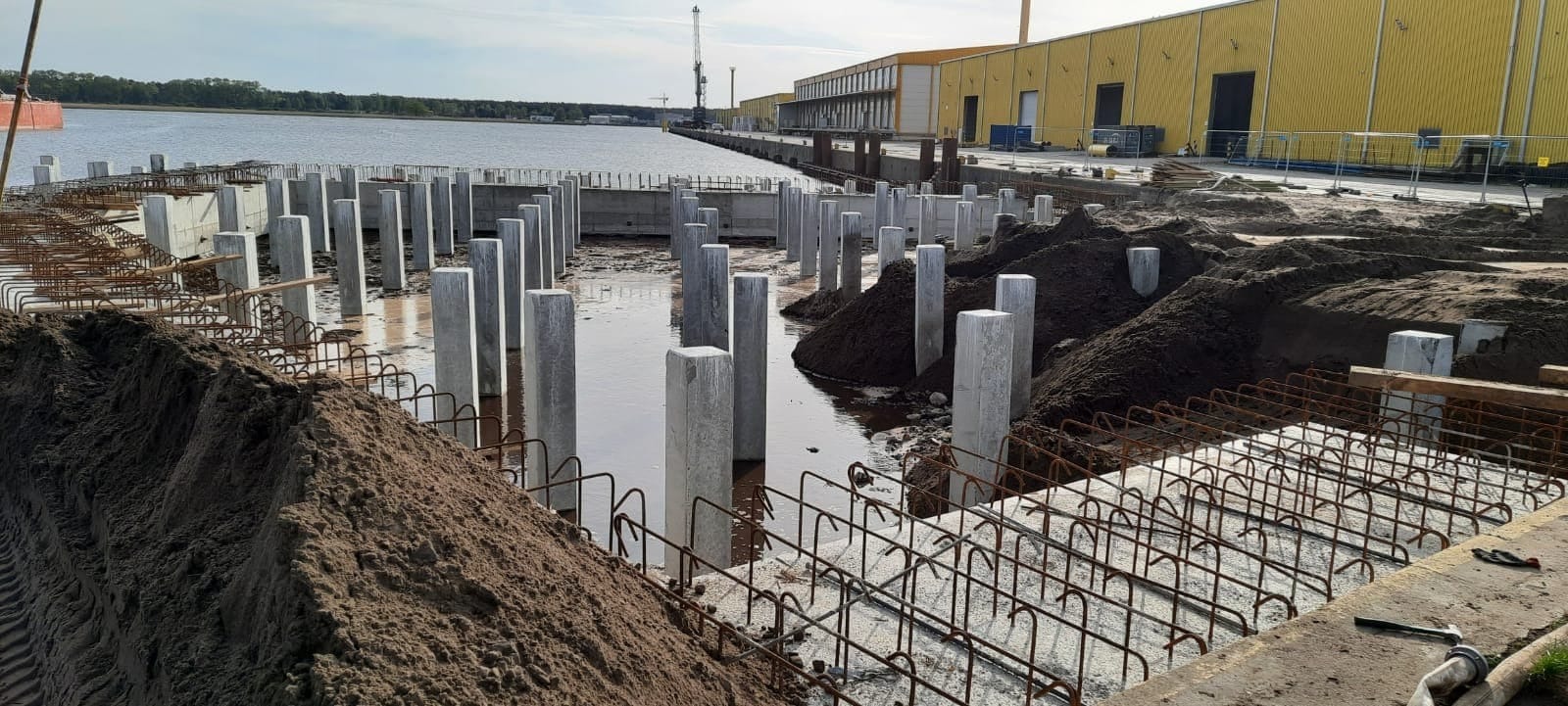 Wykonywanie żelbetowej konstrukcji nabrzeża z widocznymi pionowymi słupami i zbrojeniem w Świnoujściu, częściowo zalane wodą, z hałdami ziemi w tle.