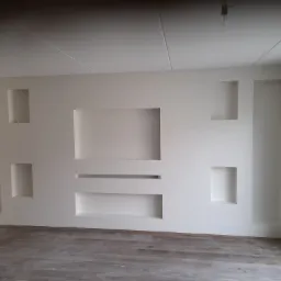 Biała ściana z wnękami o różnych kształtach i rozmiarach, minimalistyczny design, podłoga z paneli imitujących drewno, surowa żarówka na suficie.