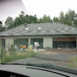 Nowo wybudowany dom jednorodzinny z szarą elewacją i ciemną dachówką, widoczny zza szyby samochodu, z dwoma osobami stojącymi przed wejściem.