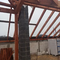 Wznoszenie komina z szarych bloczków betonowych na tle konstrukcji więźby dachowej z impregnowanego drewna, widoczne białe bloczki Ytong i drewniane deski na podmokłym podłożu.