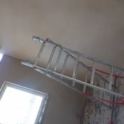 Świeżo położone tynki maszynowe na ścianach i suficie w pomieszczeniu z oknem, widoczna drabina aluminiowa z czerwonymi taśmami zabezpieczającymi.