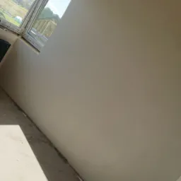 Wnętrze w Świdwinie w trakcie prac wykończeniowych, widoczna świeżo położona gładź na ścianie w jasnym odcieniu, okno z widokiem na zieleń i fragment surowej podłogi.