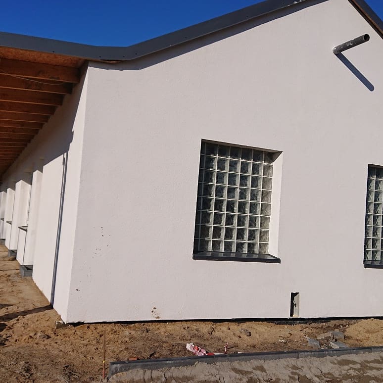 Boczny widok budynku z białym tynkiem, oknami z luksferów i szarą obróbką blacharską dachu, teren wokół budynku w stanie surowym.
