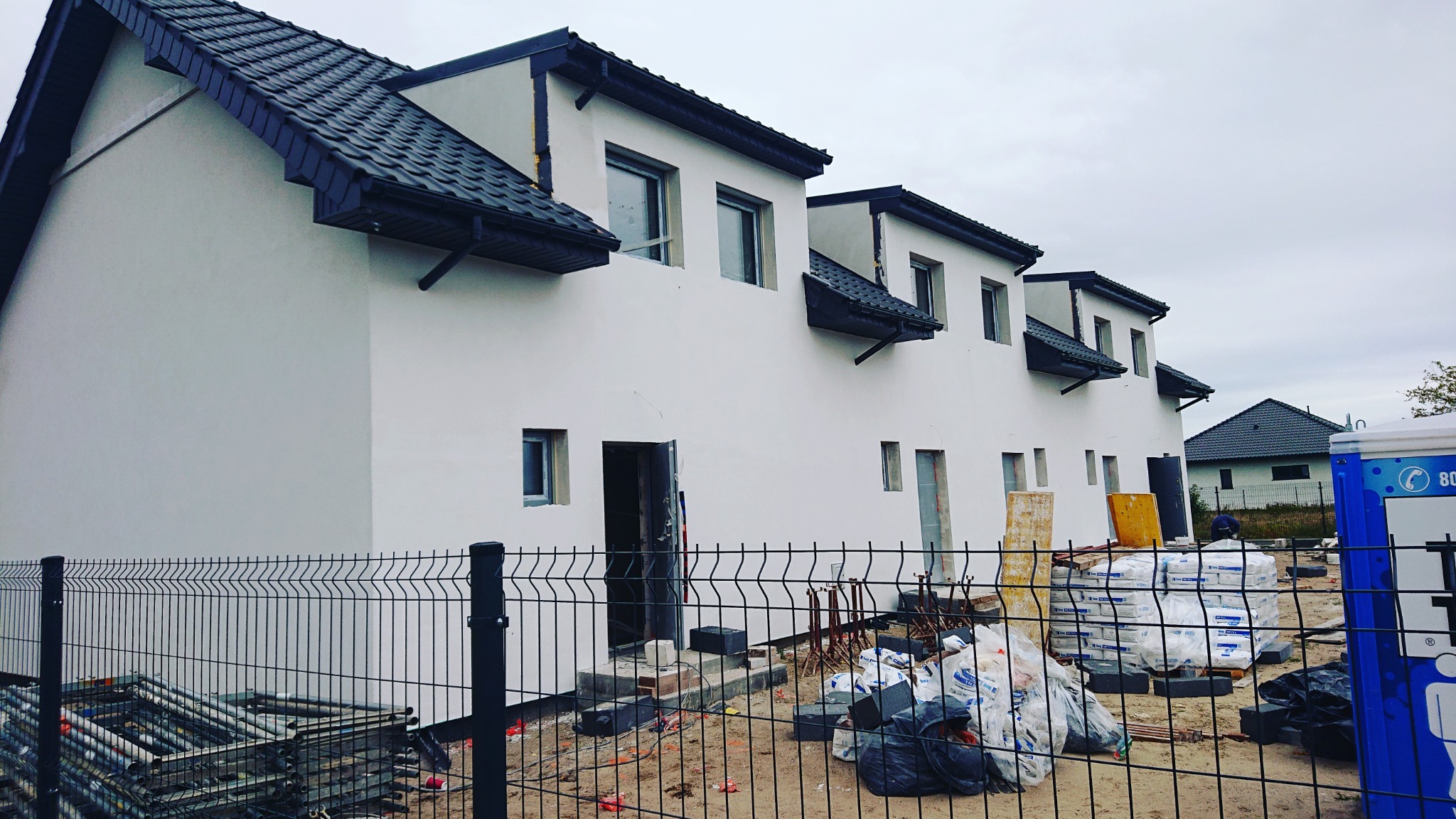 Nowo wybudowane, szeregowe domy z białymi elewacjami i grafitowym dachem, widoczne przez ogrodzenie budowlane z porozrzucanymi materiałami budowlanymi.