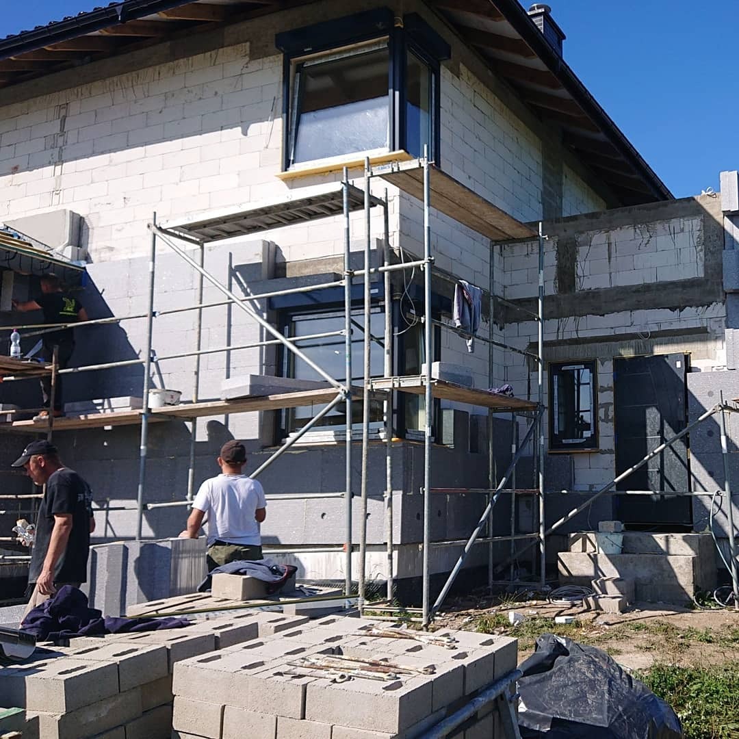 Budowa domu z białych bloczków: widoczne rusztowania, częściowo ocieplona elewacja styropianem, dwóch pracowników na rusztowaniu oraz stos bloczków na pierwszym planie.