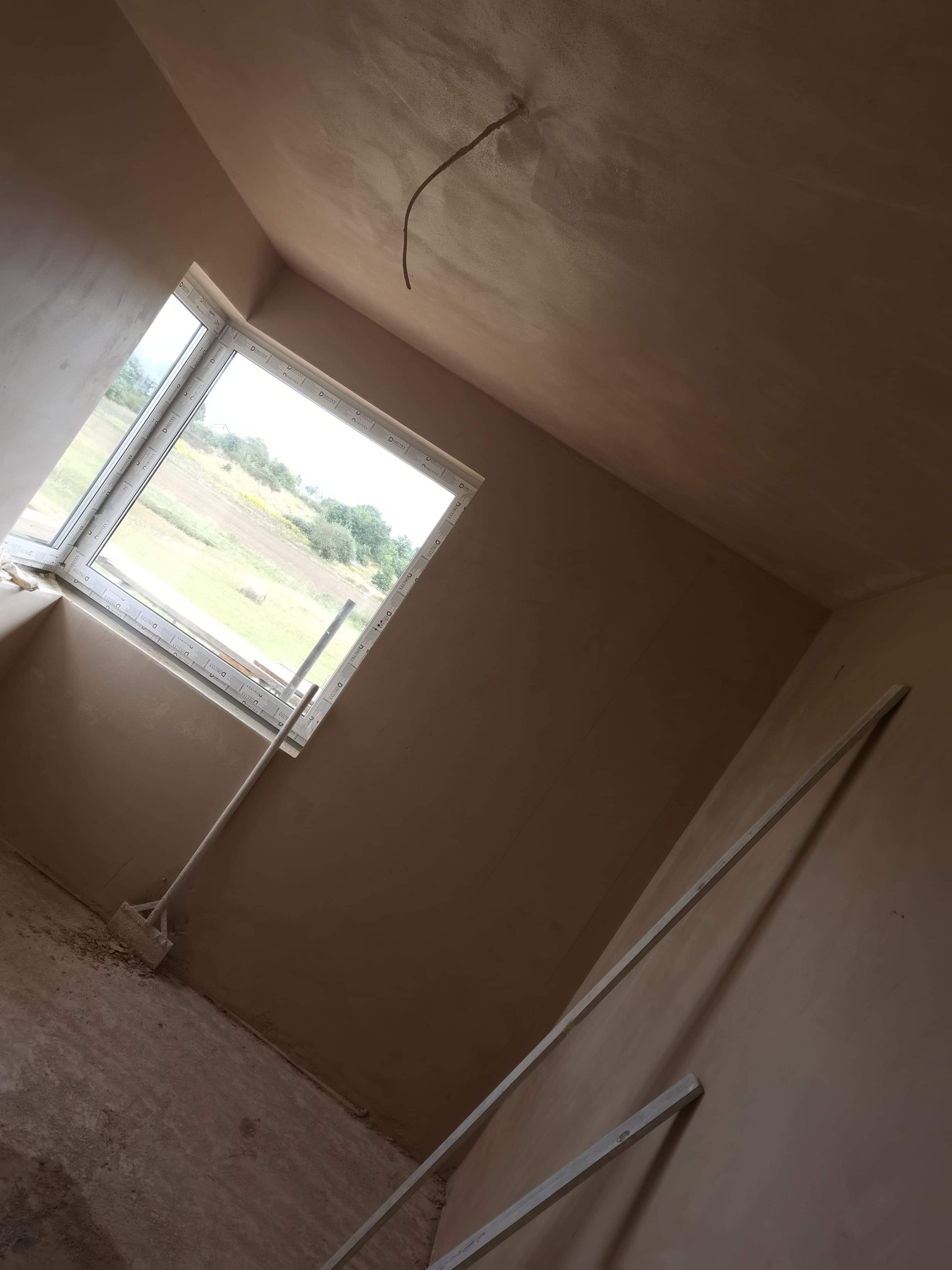 Wnętrze pokoju po nałożeniu tynku maszynowego na ściany i sufit, widoczne narzędzia: łata tynkarska oparta o ścianę i kielnia przy oknie, przez okno widok na pole i drzewa.