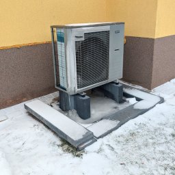 COLLTECHMAR Jeśka Mariusz - Zewnętrzna jednostka klimatyzacji przy budynku z dwukolorową elewacją, otoczona śniegiem. Widoczne betonowe podstawy i fragment okna. Zimowa sceneria.