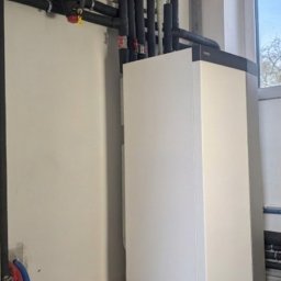 Nowoczesna, biała pompa ciepła w kotłowni z szarymi płytkami podłogowymi. Widoczne rury i zbiorniki wyrównawcze. Ujęcie z dołu, podkreślające wysokość urządzenia.