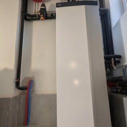 Nowoczesna, biało-szara pompa ciepła Nibe w kotłowni, z widocznymi rurami i zbiornikami wyrównawczymi. Szare płytki na podłodze i ścianach.