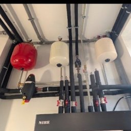 Instalacja NIBE z czerwonym zbiornikiem wyrównawczym i rurami w izolacji, zamontowana pod sufitem w jasnym pomieszczeniu. Widoczne zawory i elementy montażowe.