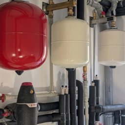 Instalacja z czerwonym i białymi zbiornikami ciśnieniowymi, rurami w izolacji i zaworami, zamontowana na białej ścianie. Widoczne manometry i elementy mocujące.