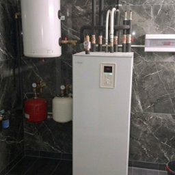 COLLTECHMAR Jeśka Mariusz - Biała pompa ciepła Nibe z licznymi rurami i zbiornikami w pomieszczeniu wykończonym płytkami imitującymi marmur. Widoczne elementy instalacji i panel sterowania.