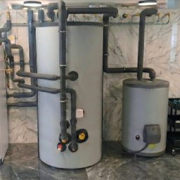 COLLTECHMAR Jeśka Mariusz - Zestaw urządzeń grzewczych: biała pompa ciepła, dwa szare zbiorniki, rury w izolacji, narzędzia na ścianie z marmurowym wzorem. Instalacja w pomieszczeniu.