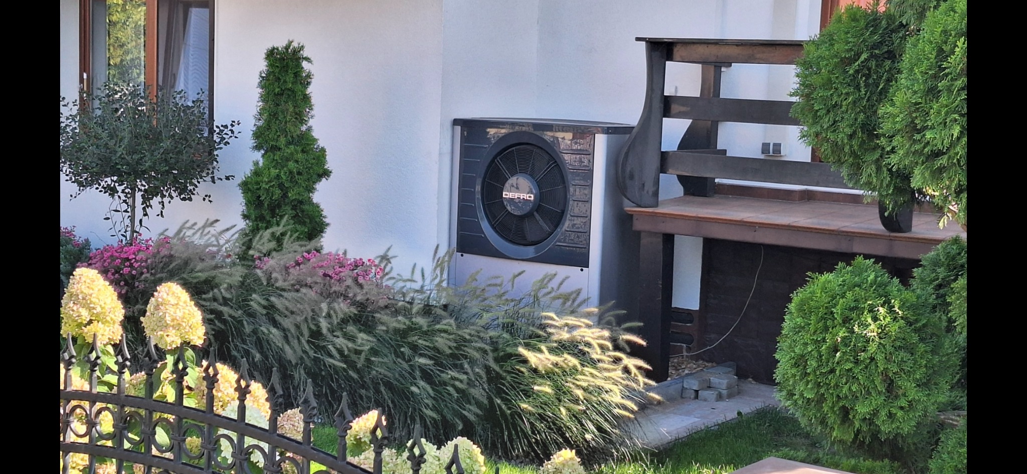 Pompa ciepła marki Defro zamontowana na białej ścianie budynku, otoczona ozdobnymi krzewami i kwiatami, z widocznym fragmentem drewnianego tarasu obok.