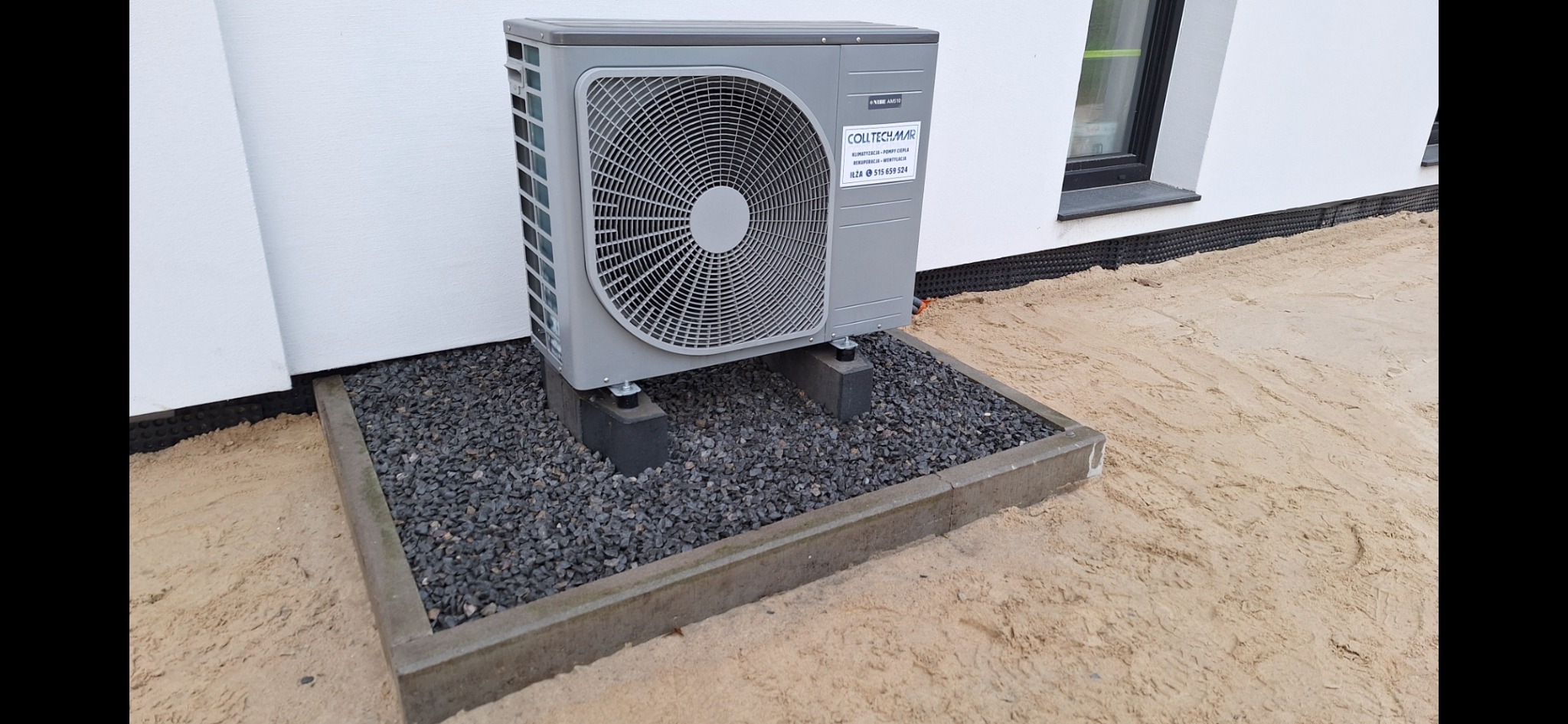 Szara pompa ciepła NIBE AMS 10 zamontowana na betonowych bloczkach, otoczona kamieniami na piaskowym podłożu przy białej ścianie budynku z widocznym oknem.