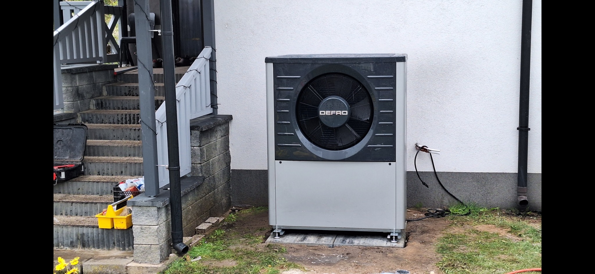 Zamontowana pompa ciepła marki DEFRO na zewnątrz budynku, widoczne podłączenia elektryczne i rura spustowa, obok schody z narzędziami.
