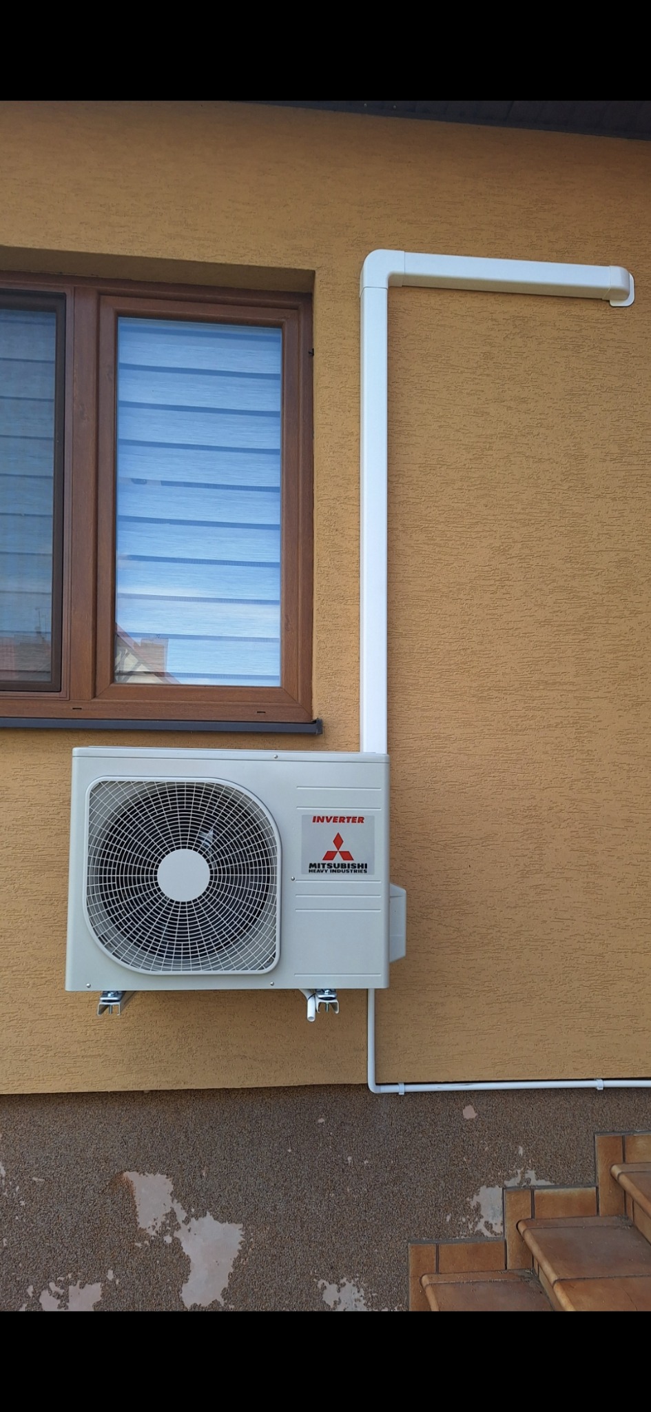 Zewnętrzna jednostka klimatyzacji Mitsubishi Inverter zamontowana na elewacji budynku z brązowym oknem i białymi rurami odprowadzającymi skropliny.