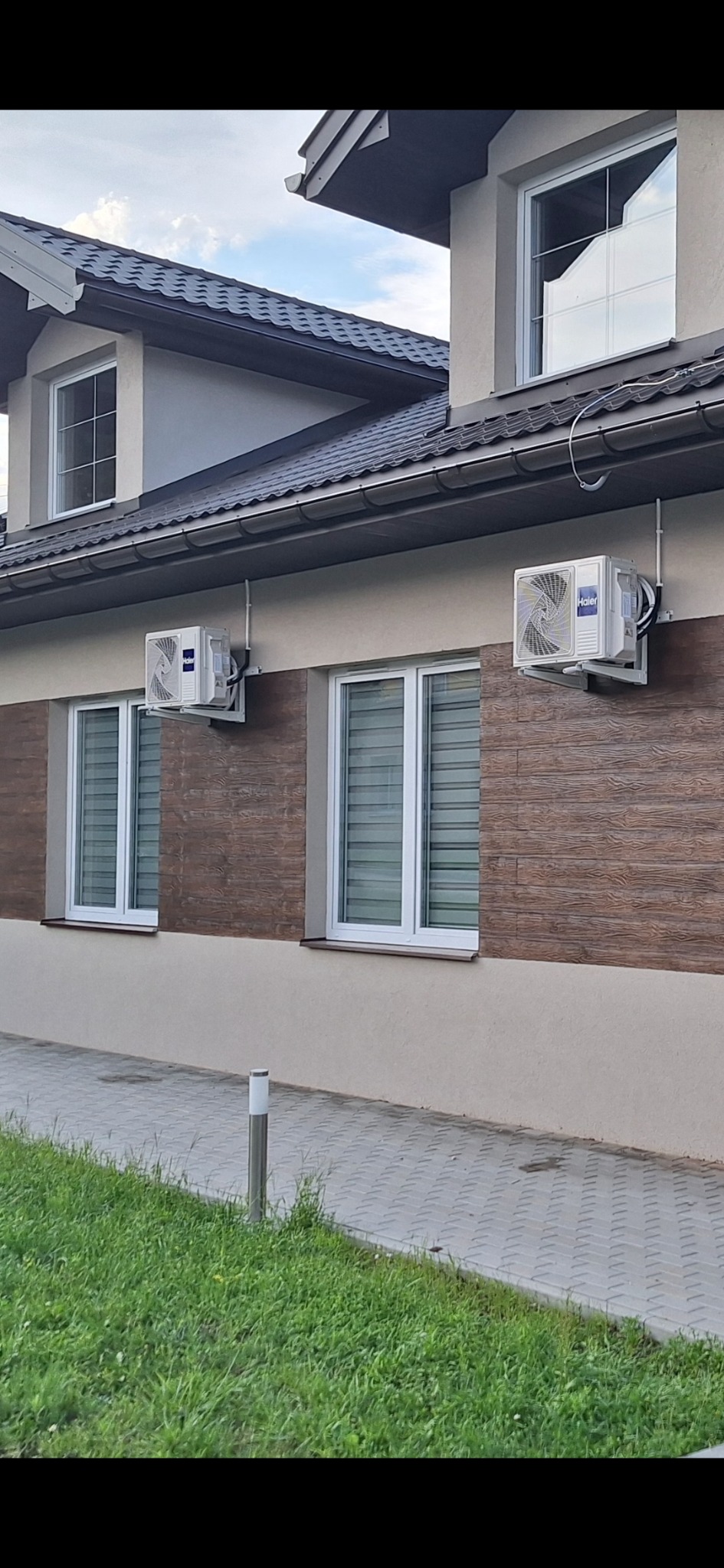 Dwa białe klimatyzatory Haier zamontowane na brązowej elewacji budynku mieszkalnego, widoczne okna z żaluzjami, trawnik i chodnik z kostki.