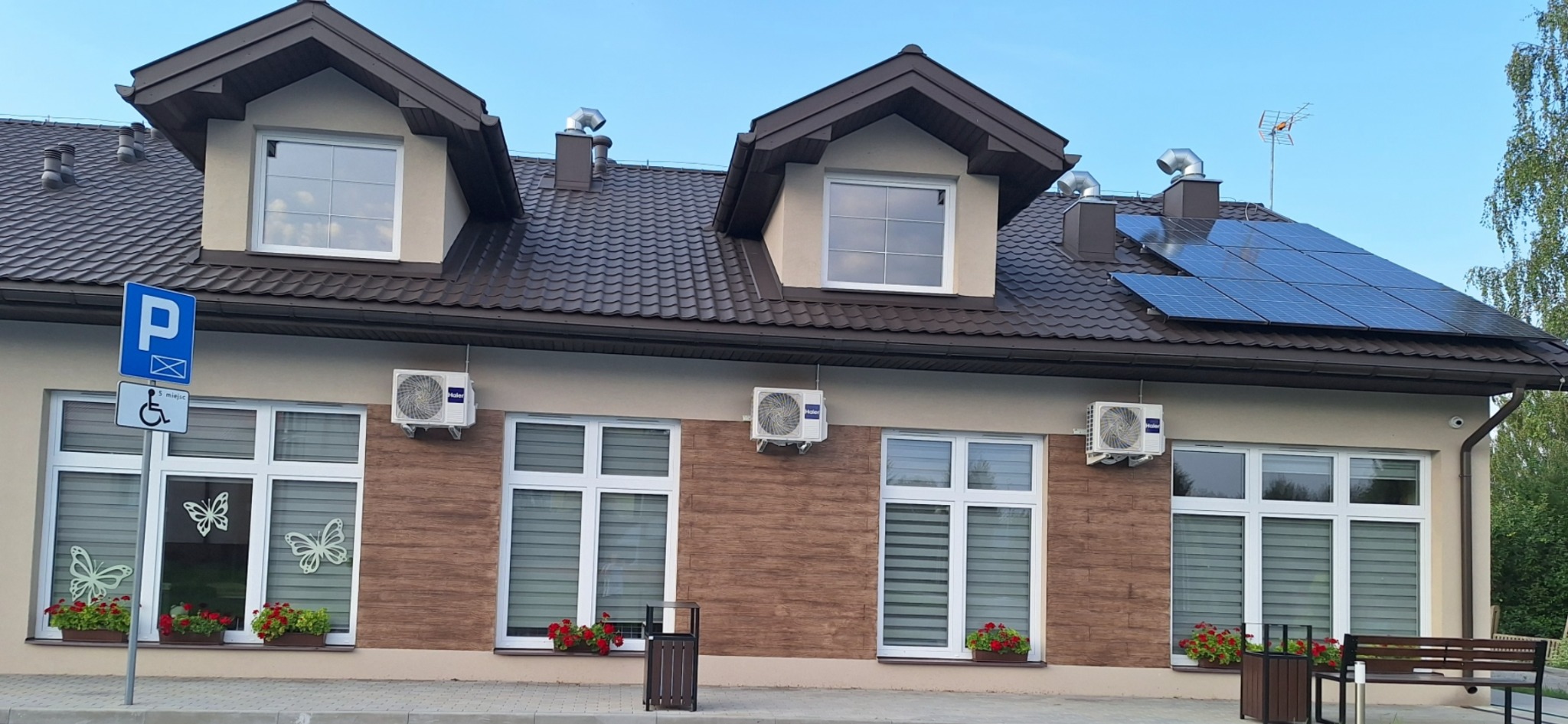 Budynek z brązowym dachem, panelami słonecznymi na dachu, klimatyzatorami na ścianie, oknami z dekoracjami motyli i skrzynkami z czerwonymi kwiatami, obok znak parkingu dla niepełnosprawnych.