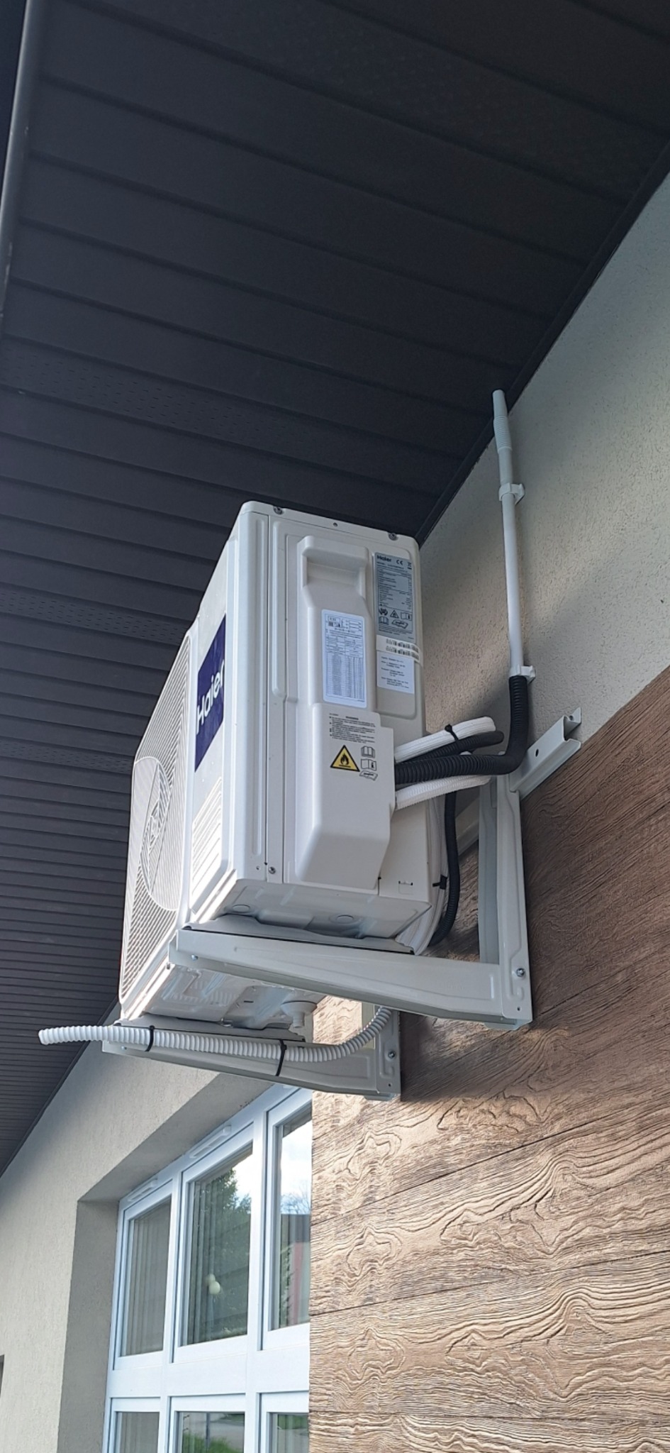 Zewnętrzna jednostka klimatyzacji Haier zamontowana na ścianie budynku z dekoracyjną okładziną imitującą drewno, widoczne rury i przewody podłączone do urządzenia.