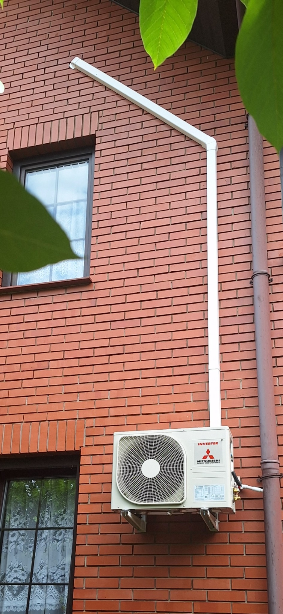 Biała jednostka zewnętrzna klimatyzacji Mitsubishi Inverter zamontowana na ceglanej ścianie budynku, z widocznym orurowaniem i elementami instalacji odprowadzającej skropliny.