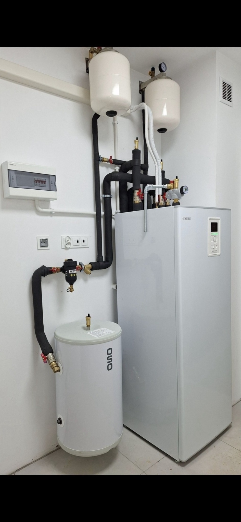 Biała pompa ciepła Nibe z zbiornikiem OSO, rury w izolacji, manometry i zawory w kotłowni, widoczna skrzynka elektryczna na ścianie.