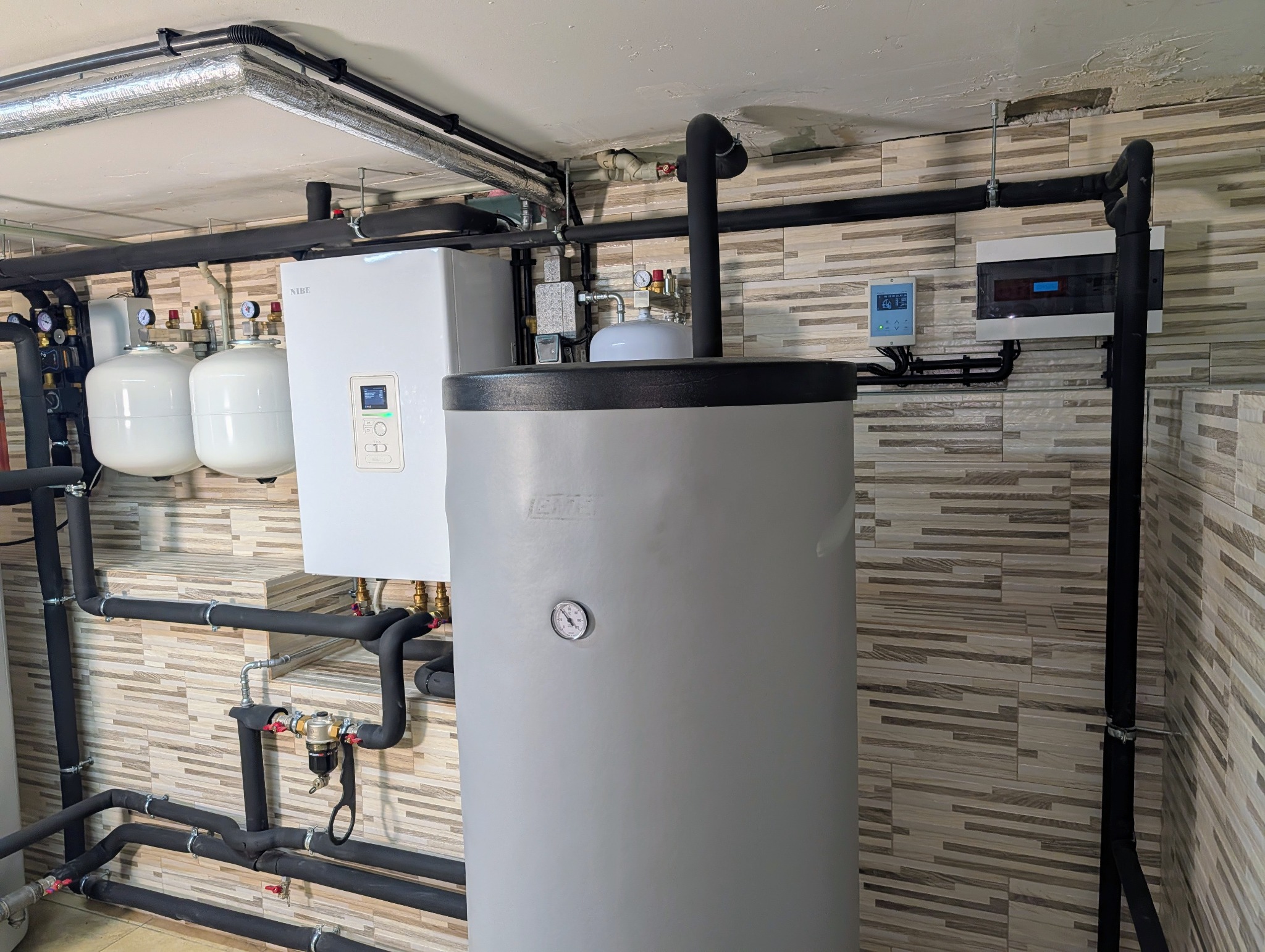Zestaw urządzeń grzewczych Nibe: pompa ciepła, zbiornik buforowy i naczynia wzbiorcze. Czarna instalacja rurowa na tle ściany z płytek imitujących drewno.