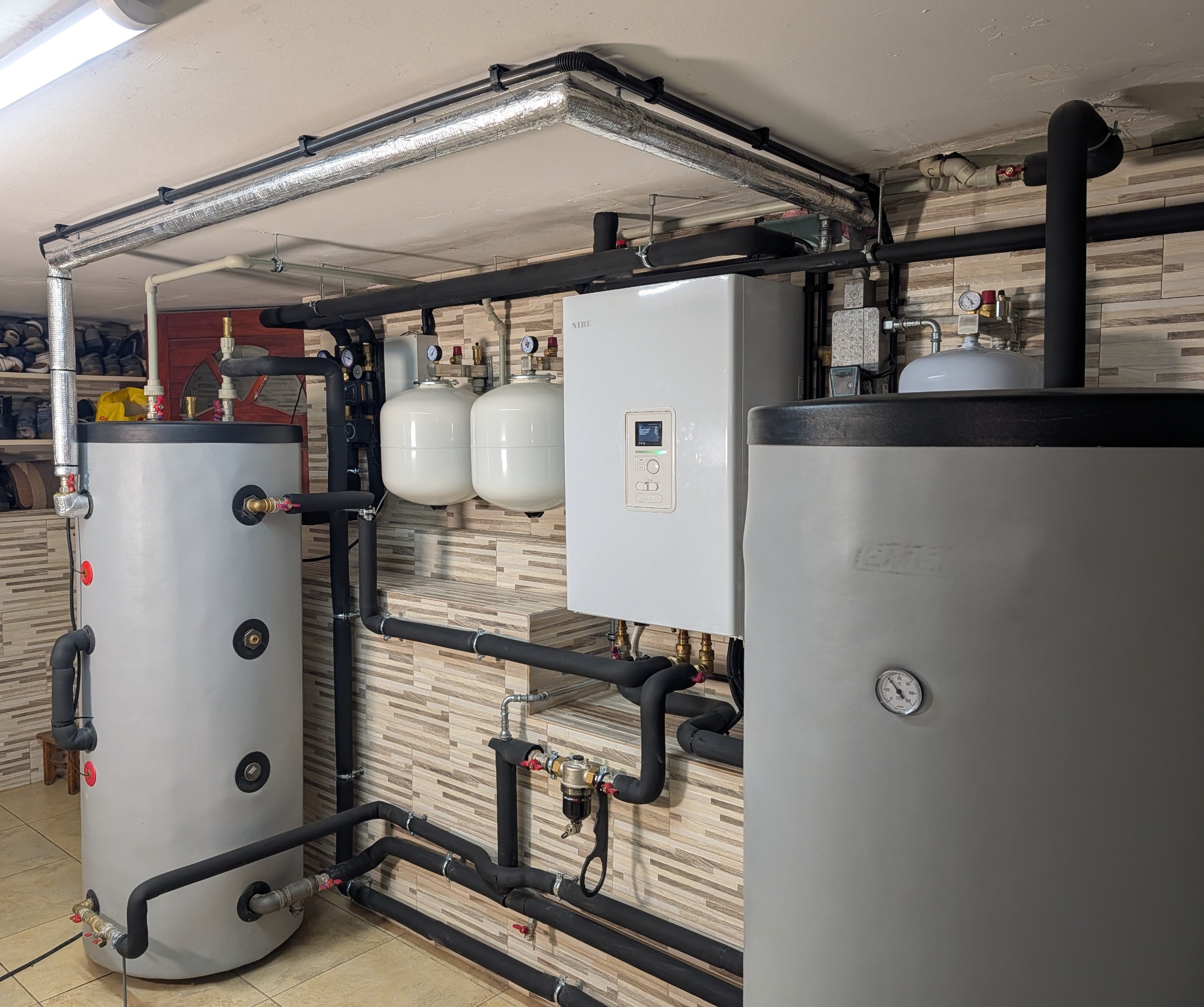 Złożona instalacja grzewcza w kotłowni: dwa zbiorniki, biały piec NIBE z wyświetlaczem, rury w izolacji, manometry. Całość na tle ściany z płytek imitujących drewno.