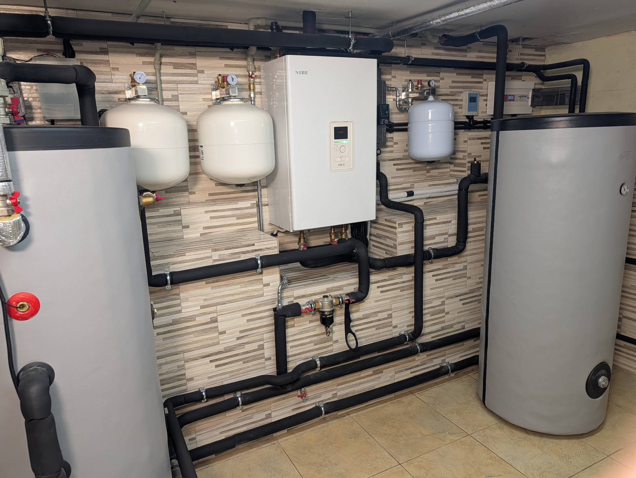 Zmontowana instalacja z pompą ciepła Nibe SHB 20, zbiornikami buforowymi i rurami w izolacji, na tle ściany z płytek imitujących drewno.