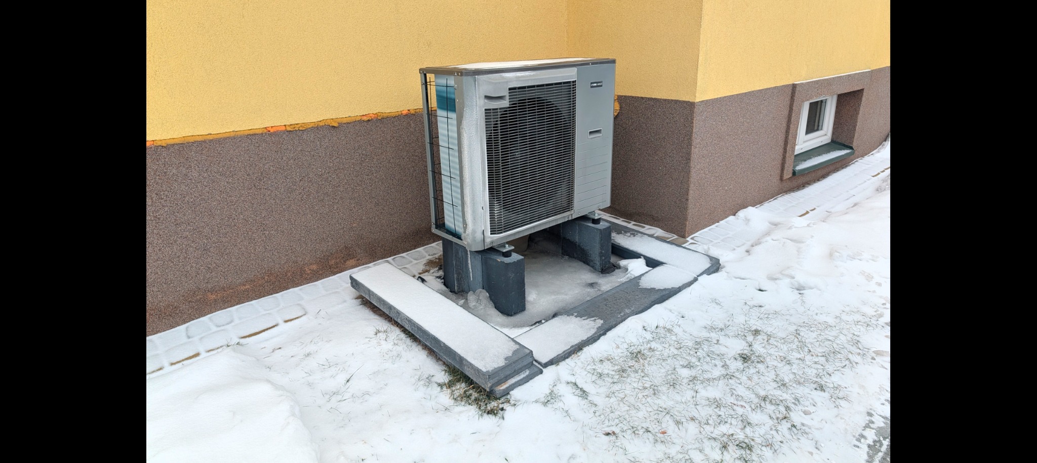 Zewnętrzna jednostka klimatyzacji przy budynku z dwukolorową elewacją, otoczona śniegiem. Widoczne betonowe podstawy i fragment okna. Zimowa sceneria.