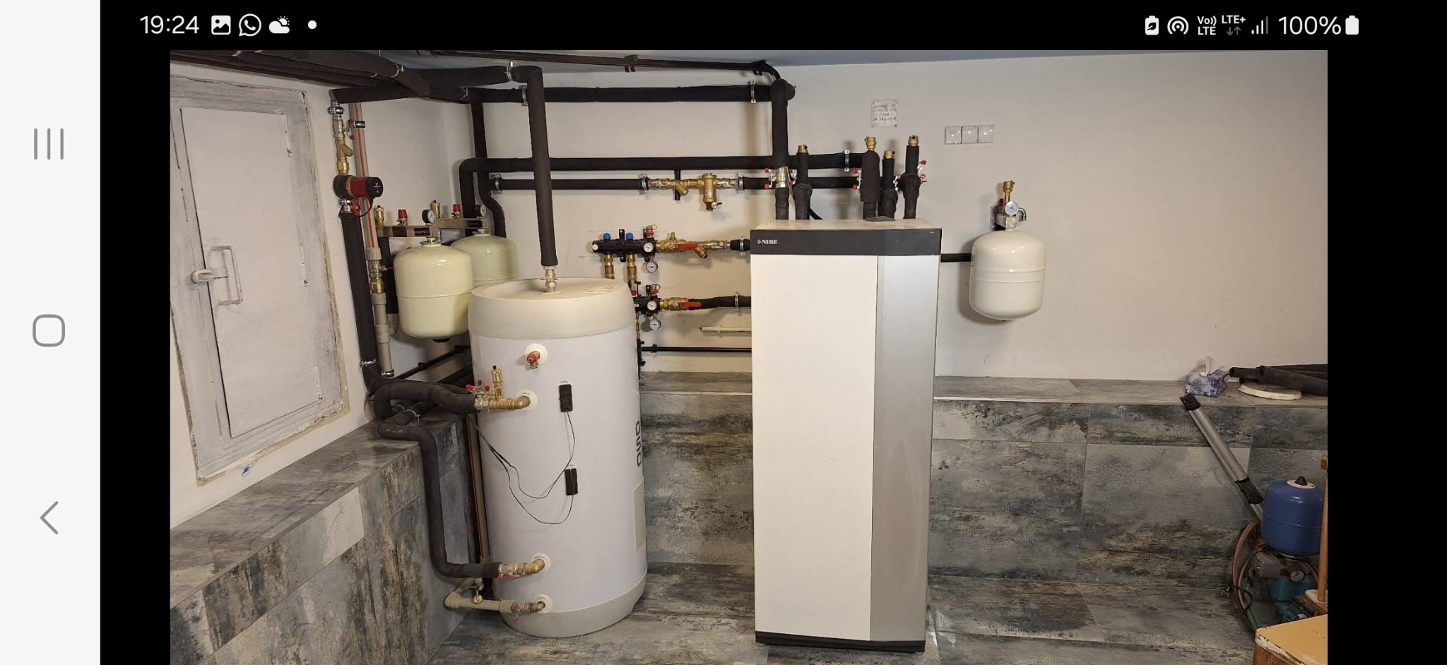 Złożona instalacja z pompą ciepła Nibe, zbiornikiem Atlantic i rurami w piwnicy. Widoczne liczne zawory i elementy montażowe. Szare płytki na podłodze.