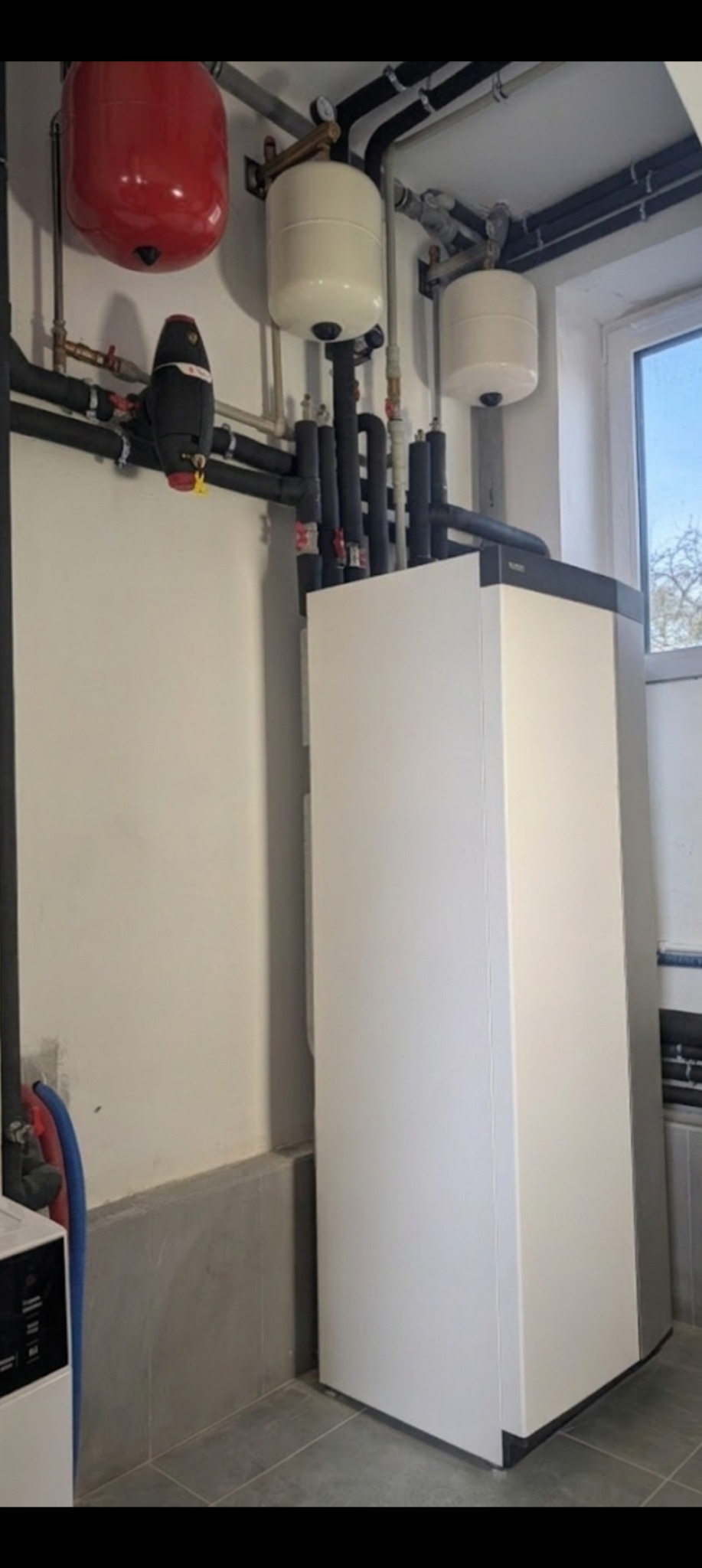 Nowoczesna, biała pompa ciepła w kotłowni z szarymi płytkami podłogowymi. Widoczne rury i zbiorniki wyrównawcze. Ujęcie z dołu, podkreślające wysokość urządzenia.