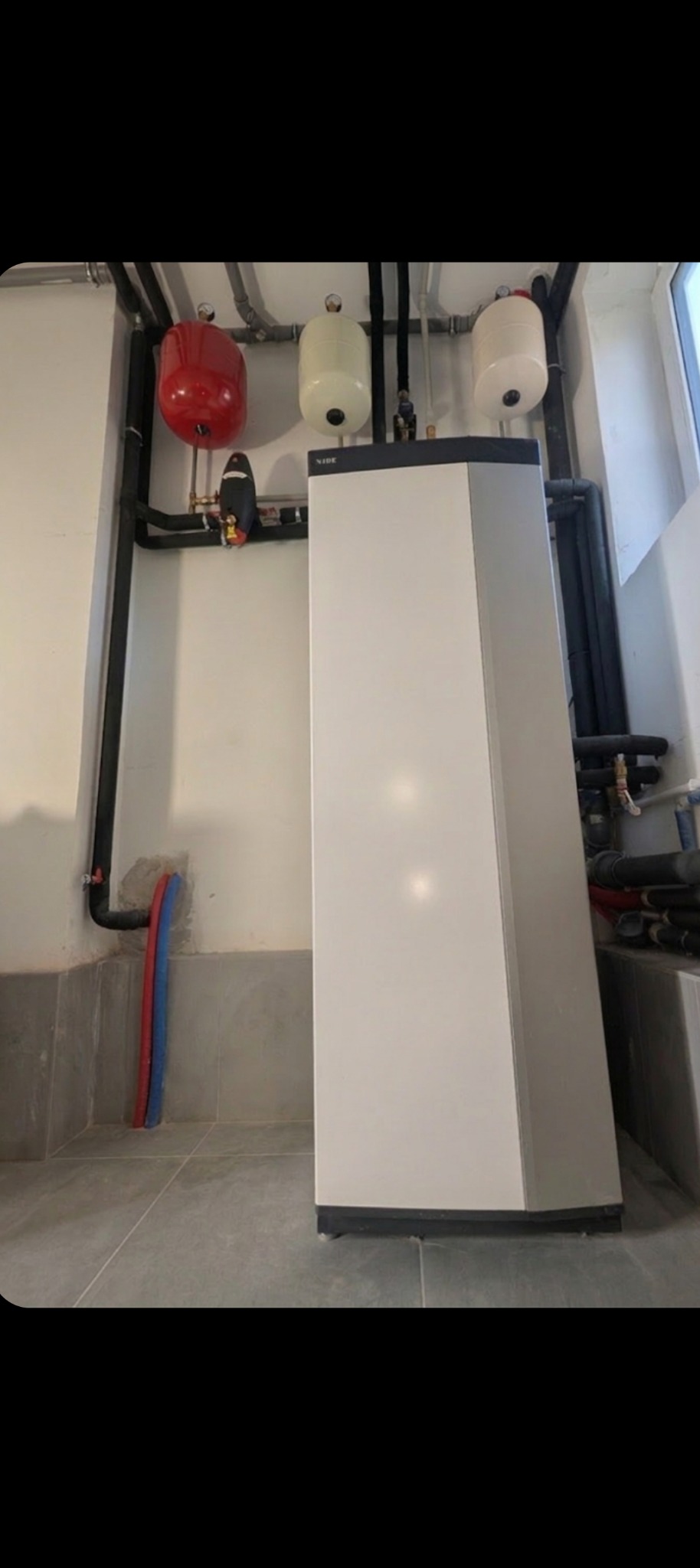 Nowoczesna, biało-szara pompa ciepła Nibe w kotłowni, z widocznymi rurami i zbiornikami wyrównawczymi. Szare płytki na podłodze i ścianach.