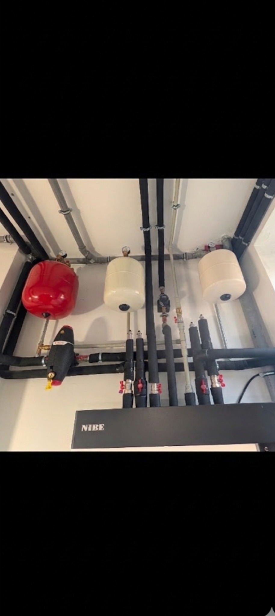 Instalacja NIBE z czerwonym zbiornikiem wyrównawczym i rurami w izolacji, zamontowana pod sufitem w jasnym pomieszczeniu. Widoczne zawory i elementy montażowe.