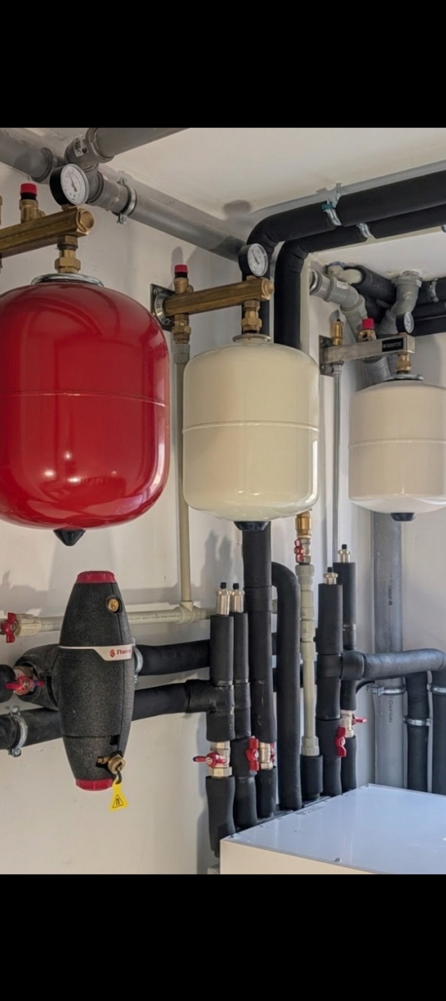 Instalacja z czerwonym i białymi zbiornikami ciśnieniowymi, rurami w izolacji i zaworami, zamontowana na białej ścianie. Widoczne manometry i elementy mocujące.