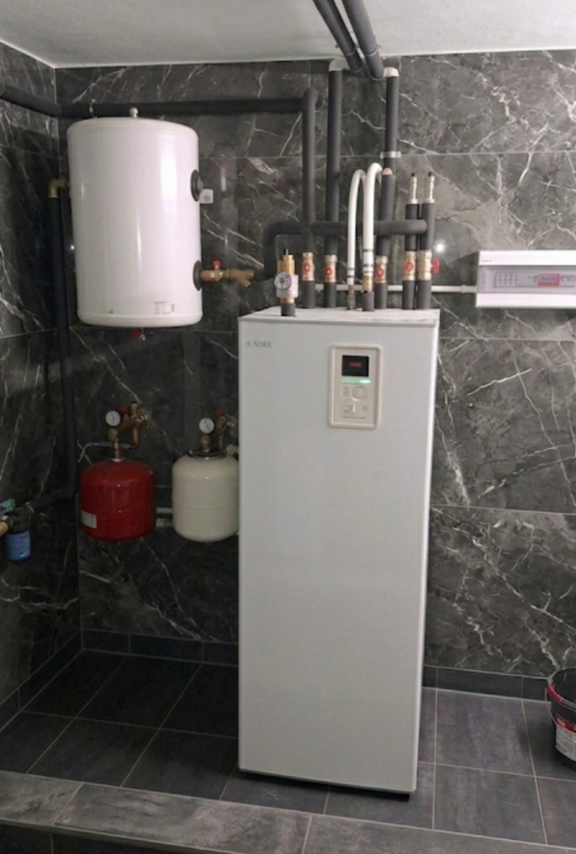 Biała pompa ciepła Nibe z licznymi rurami i zbiornikami w pomieszczeniu wykończonym płytkami imitującymi marmur. Widoczne elementy instalacji i panel sterowania.