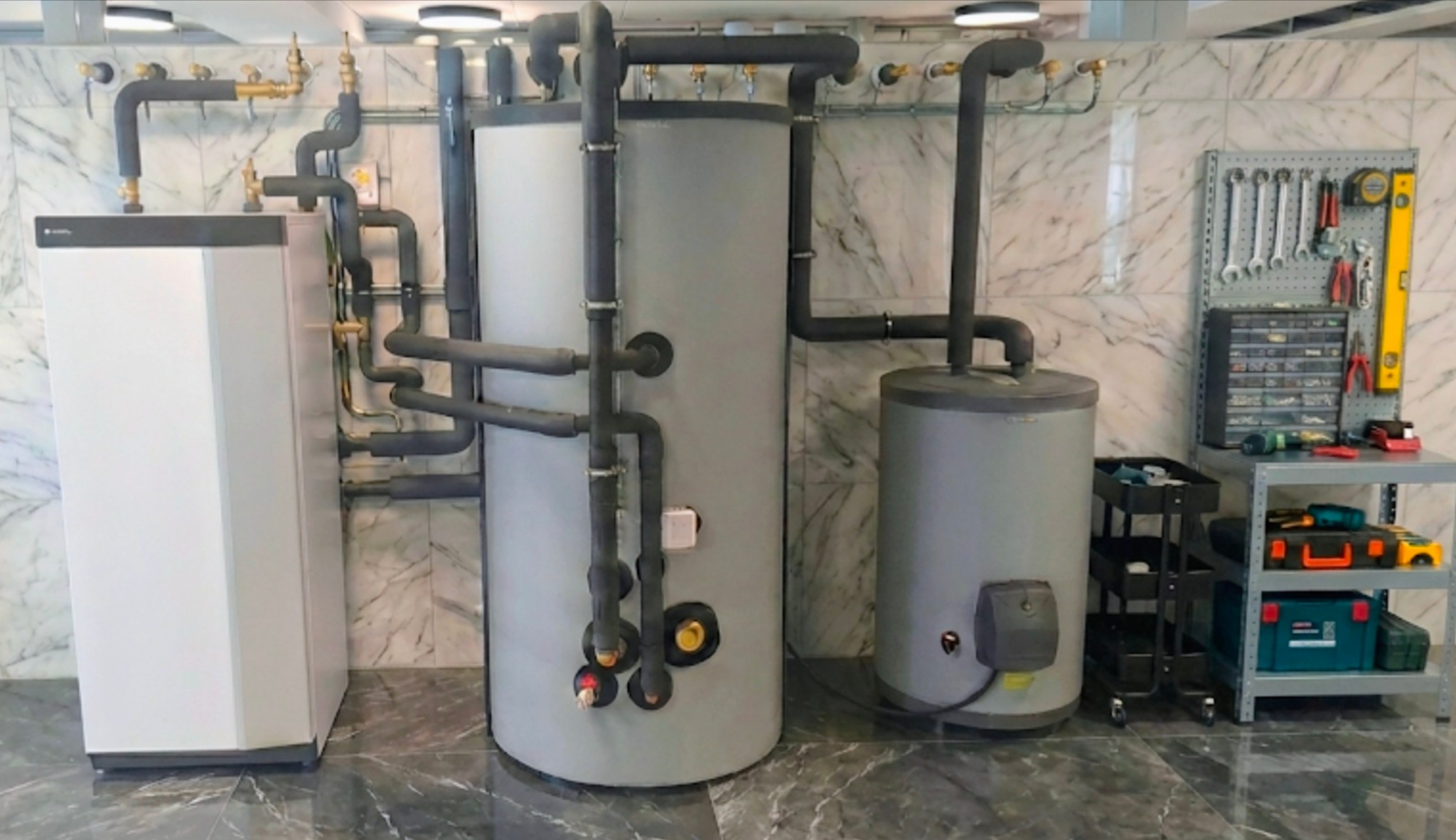 Zestaw urządzeń grzewczych: biała pompa ciepła, dwa szare zbiorniki, rury w izolacji, narzędzia na ścianie z marmurowym wzorem. Instalacja w pomieszczeniu.